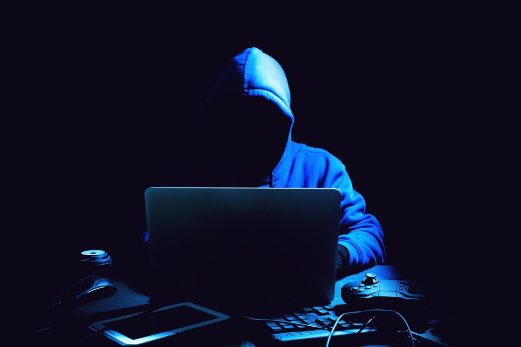 A shadowy figure in a hoodie sits at a laptop