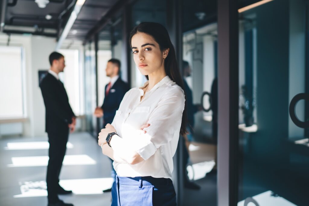 A young female businesswoman stands in an office corridor with her arms folded