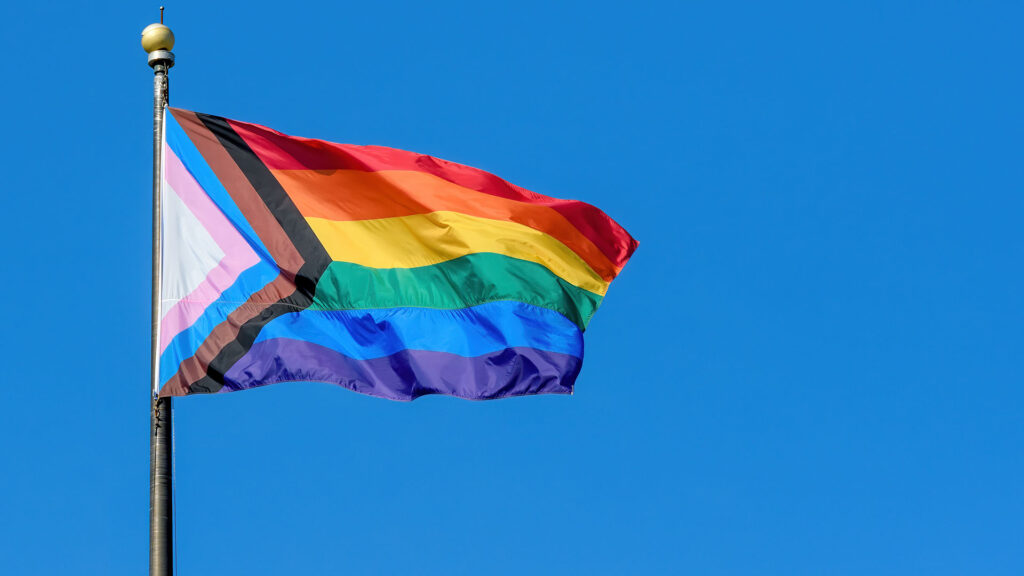 A LGBTQ+ rainbow progress pride flag flaps from a flagpole in a brisk breeze. Blue sky behind.