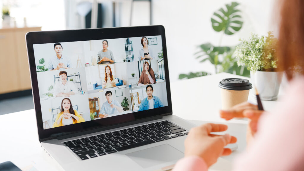 A photograph of a laptop with a teleconference on screen taken over the shoulder of a remote worker at home