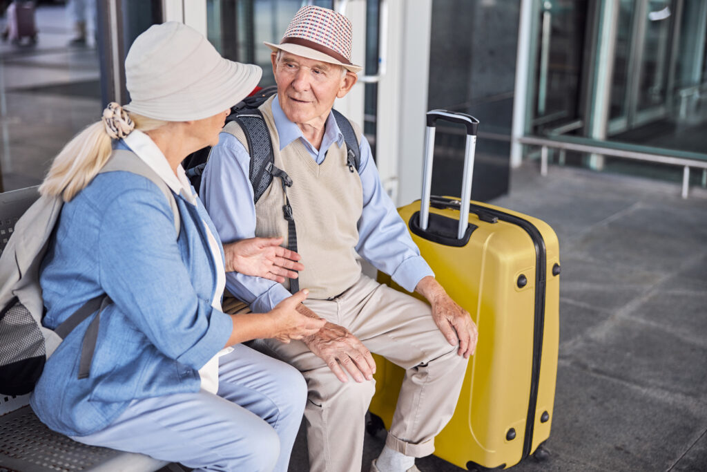 A senior couple converses on a bench next to their luggage