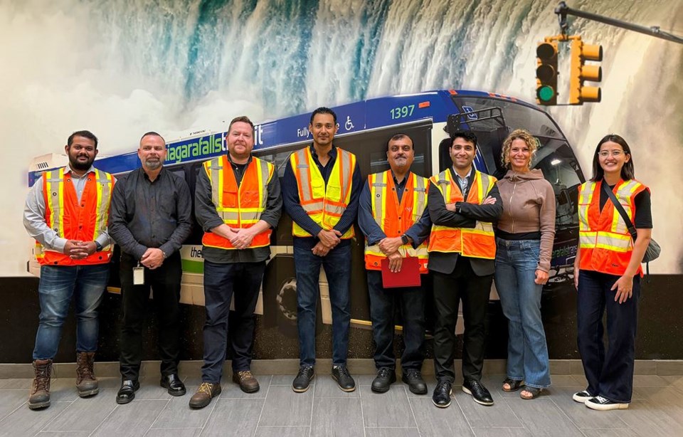 A group of workers in hi-viz vests stand in front of a bus with a backdrop of Niagara Falls behind