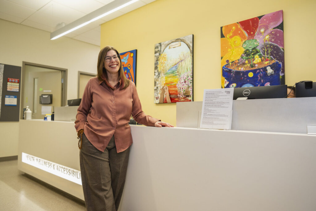 Dr. Karen Csoli, Director, Health, Wellness & Accessibility Services, Niagara College Welland Campus. A white woman with blonde hair and spectacles wearing an orange shirt and tan pants.