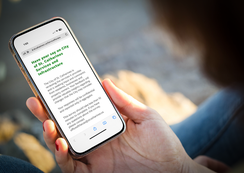 A woman's hand holds a cellphone displaying the City of St. Catharines core service review page