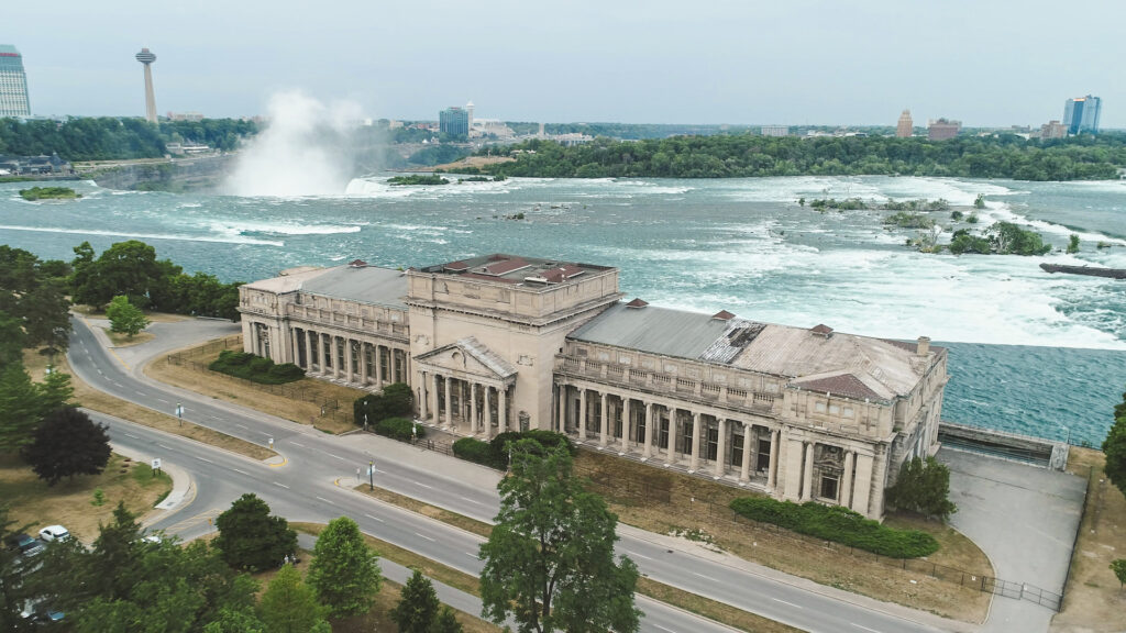 An aerial drone photograph of the Toronto Power Generating Station building, a neoclassical building with two wings in a disused state, with Niagara Falls in the background