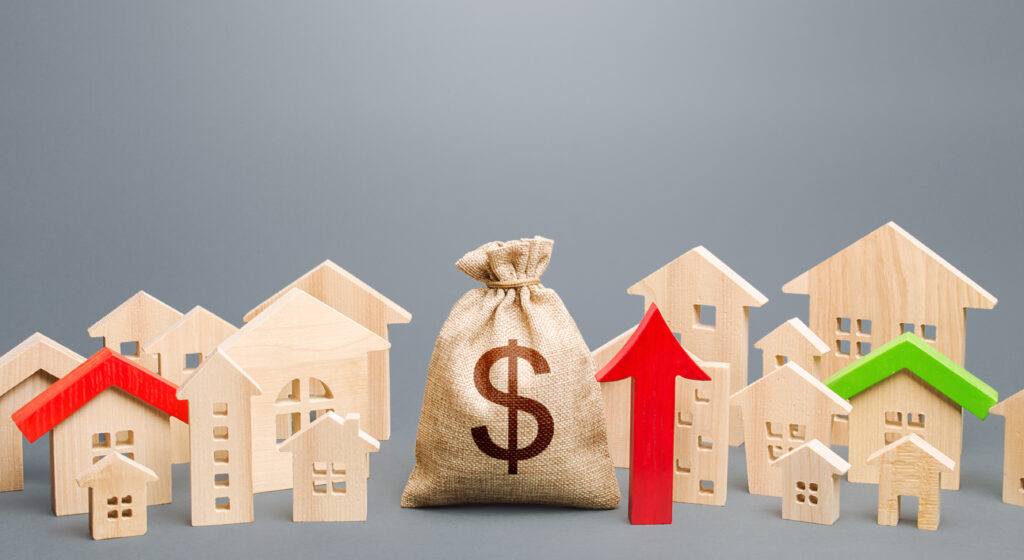 A sack with a dollar sign printed on it sits next to a red arrow pointing upwards in the middle of a group of wooden model houses