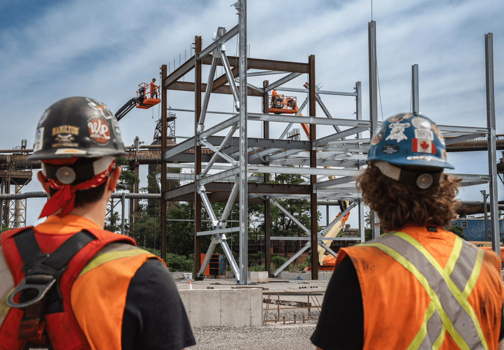 Two workers in hard hats and hi-viz vests look on as the steel frame of a building is assembled