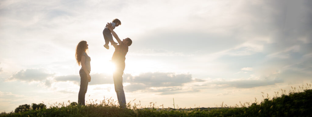 A family silhouetted against the sky. The father is holding a young child in the air while the mother looks on.