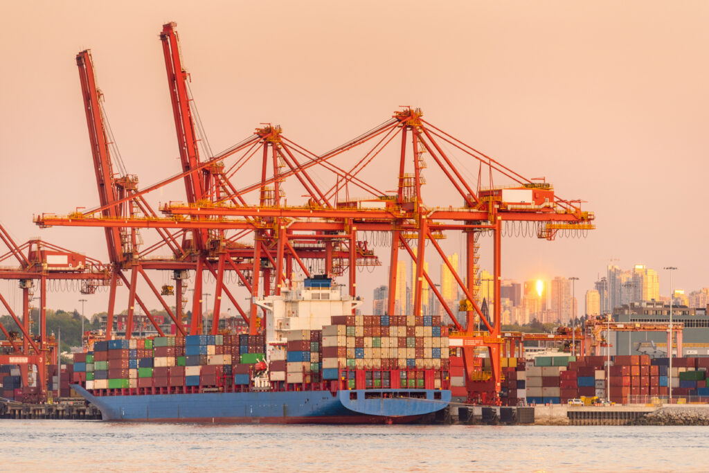 A container ship docked at the Port of Vancouver with a row of orange cranes behind it and the city skyline in the background