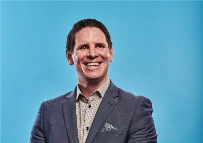 Pharmacist Sean Simpson. A smiling white man with dark hair wearing a grey suit jacket and beige checked button-up shirt.