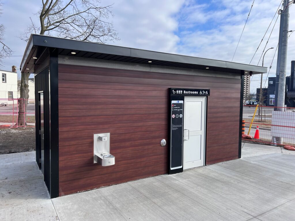 The self-cleaning washroom in St. Catharines. A brown, box-shaped structure with a white door and a drinking fountain on the exterior wall.