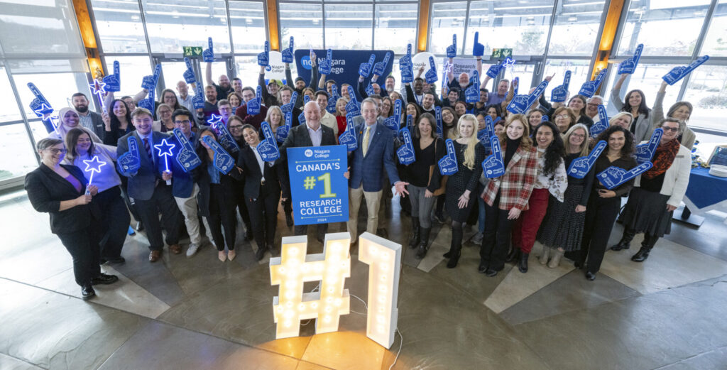 Niagara College President Sean Kennedy (holding sign) and college employees, including members of the Research and Innovation team, celebrate NC’s No. 1 research ranking on December 5 at the Daniel J. Patterson Campus in Niagara-on-the-Lake.