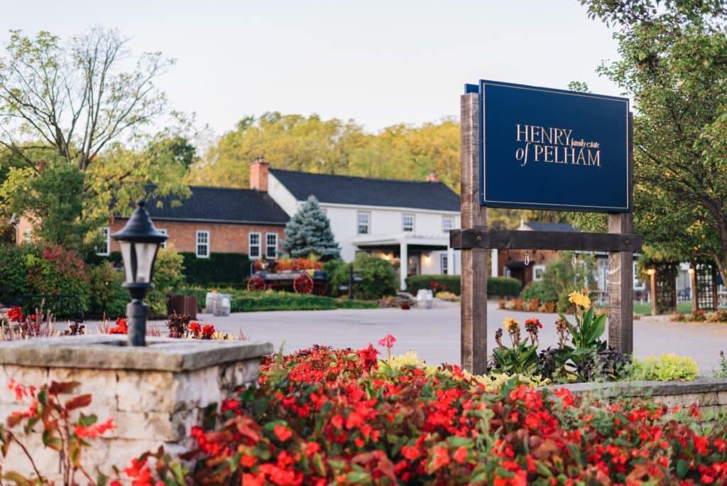 The sign at Henry of Pelham estate winery. A blue sign in a flowerbed with traditional buildings in the background.