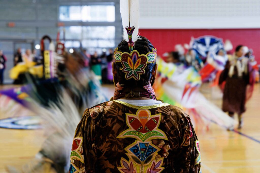 A person in traditional Indigenous clothing watches performers at a Pow Wow in Brock University's Ian Beddis gymnasium